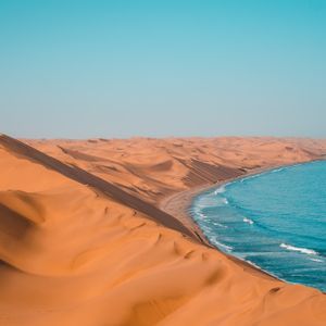 Weite orangefarbene Sanddünen treffen auf den blauen Ozean und bilden eine Küstenlinie unter klarem Himmel.