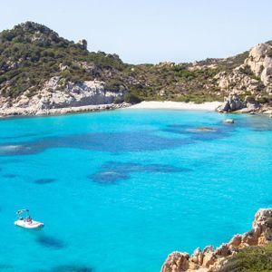 Numerose barche e barche a vela sono ormeggiate in una baia dalle acque cristalline turchesi, circondata da colline rocciose e una spiaggia sabbiosa.