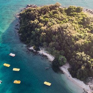 Vista aerea di una piccola isola ricoperta di alberi in acque turchesi, con diversi pedalò gialli che galleggiano nelle vicinanze.