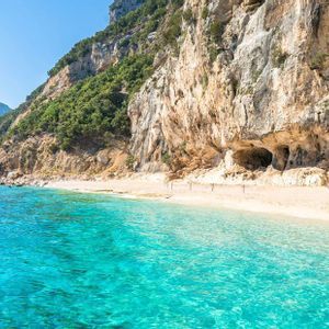 Acque turchesi lambiscono una spiaggia sabbiosa e appartata ai piedi di un'imponente scogliera frastagliata sotto un cielo limpido.