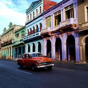 Un'auto d'epoca arancione percorre una strada accanto a colorati edifici coloniali con portici ad arco sotto un cielo blu.