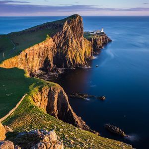 Un phare blanc se dresse sur une péninsule rocheuse et herbeuse qui s'avance dans la mer bleue et calme, baigné par la lumière chaude du coucher de soleil.
