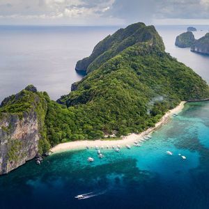 Aerial view of a lush, rocky island featuring a white sand beach with many boats moored in the surrounding turquoise water.