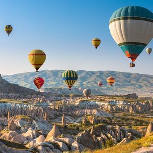 Globos aerostáticos de colores vuelan sobre un vasto paisaje rocoso con formaciones cónicas bajo un cielo despejado.
