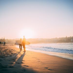 Zwei silhouettierte Surfer mit Surfbrettern stehen an einem Sandstrand und blicken bei einem goldenen Sonnenuntergang auf das Meer.