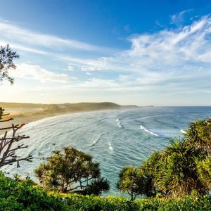An elevated view from a lush green clifftop of a sandy beach curving along the coastline next to the ocean under a blue sky.