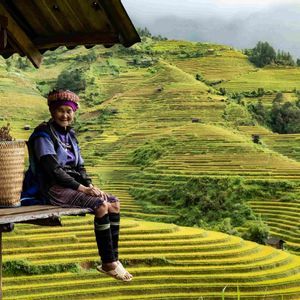 Una anciana con vestimenta tradicional se sienta sonriendo en un porche de madera con vistas a vastos arrozales en terrazas en una ladera.