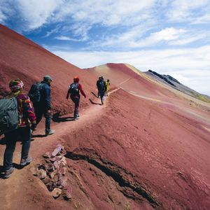 Un viaggio di gruppo WeRoad facendo trekking su uno stretto sentiero lungo il fianco di una vasta montagna rossa sotto un cielo blu parzialmente nuvoloso.