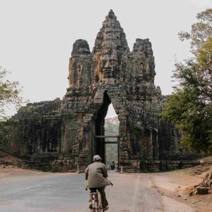 A person seen from behind rides a bicycle on a road towards a monumental ancient stone gate carved with large faces.