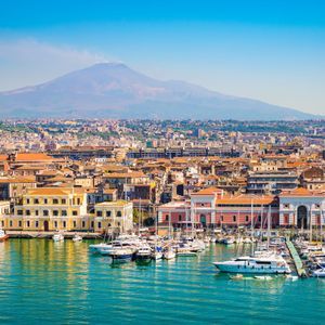 A coastal city's harbor filled with boats, with a large mountain rising in the background under a clear blue sky.