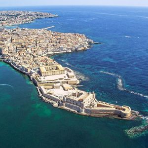 Una vista aerea di una storica città costiera e fortezza su una penisola, circondata da mare turchese e blu intenso.