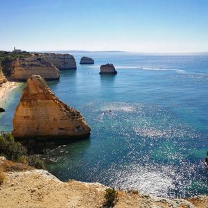 Un capo gruppo WeRoad con un cappello siede su una scogliera che si affaccia su una costa rocciosa con archi naturali sul mare e una piccola spiaggia.