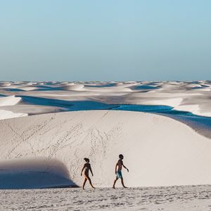 Zwei Personen gehen über weite, weiße Sanddünen, die mit blauen Lagunen gesprenkelt sind, unter einem klaren Himmel.
