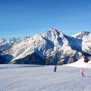 Personen beim Skifahren auf einer breiten, verschneiten Piste mit einer riesigen Bergkette und einer Skihütte im Hintergrund unter klarem blauem Himmel.