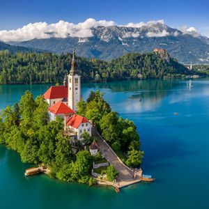 A white church with red roofs sits on a tree-covered island in a turquoise lake, with steep mountains in the background.