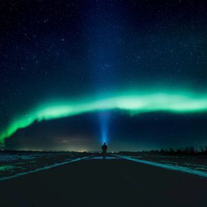A person stands on a dark road, shining a flashlight up at the green aurora borealis under a starry night sky.