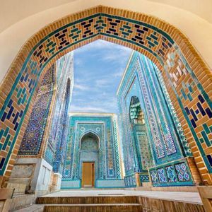 Ein Blick durch einen großen Ziegel- und Fliesenbogen auf einen Innenhof mit Gebäuden, die mit aufwendigen blauen und türkisen Mosaiken bedeckt sind.