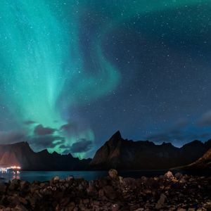 L'aurora boreale verde risplende in un cielo notturno stellato su un fiordo, con montagne stagliate e una costa rocciosa sottostante.