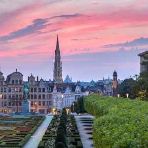 Una vista panoramica su un giardino formale verso lo skyline di una storica città europea con un'alta guglia sotto un vivace tramonto rosa.