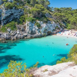 Vue aérienne d'une crique où des personnes nagent dans une eau turquoise claire et se détendent sur une plage de sable blanc entourée de falaises rocheuses et d'arbres.