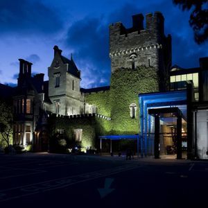 Un castello in pietra ricoperto d'edera con un ingresso moderno è illuminato da luci sotto un cielo notturno blu scuro.