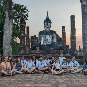 Un voyage de groupe WeRoad s'assied en tailleur pour une photo devant une grande statue de Bouddha au milieu de ruines de temples anciens au coucher du soleil.