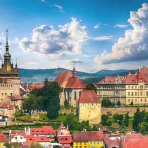 A historic town with a central clock tower and colorful buildings with red roofs, set against a backdrop of green hills and a blue sky.