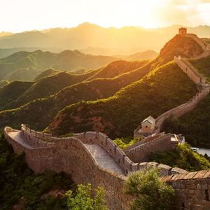 Die Chinesische Mauer schlängelt sich über sanfte grüne Berge, wobei ein Wachturm im goldenen Licht eines Sonnenuntergangs sichtbar ist.