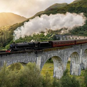 Un treno a vapore nero con carrozze rosse attraversa un viadotto in pietra curvilineo, emettendo fumo bianco in una valle verde e soleggiata.