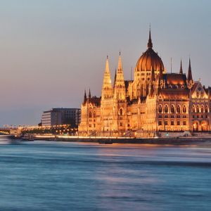 A large, ornate building with a central dome is illuminated by golden lights along a river at dusk.