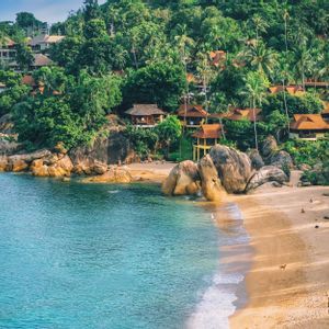 High-angle view of a sandy beach cove with turquoise water, large boulders, and villas nestled in a lush, green, palm-filled hillside.