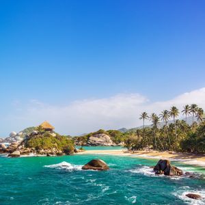 A hut with a thatched roof sits on a rocky outcrop in turquoise waters, next to a sandy beach lined with palm trees under a blue sky.
