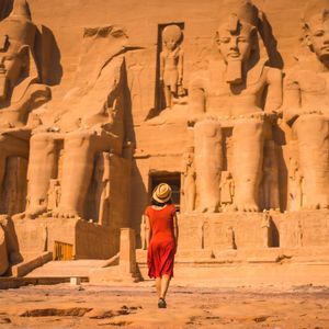 A woman in a red dress and straw hat walks towards a large ancient temple carved into a cliff with colossal seated statues.