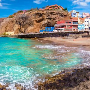 A panoramic view of a coastal village with colorful houses built into a cliff above a turquoise sea and sandy beach.