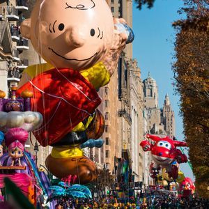Un gigantesco pallone di Charlie Brown e altri carri a tema avanzano lungo una via cittadina durante una parata sotto un cielo azzurro e limpido.