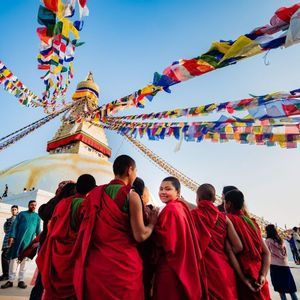 A group of people in red robes look towards a large white stupa with colorful prayer flags strung from its spire against a blue sky.
