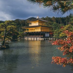 A golden Japanese temple stands by a reflective pond, surrounded by autumn trees and forested hills under a cloudy sky.
