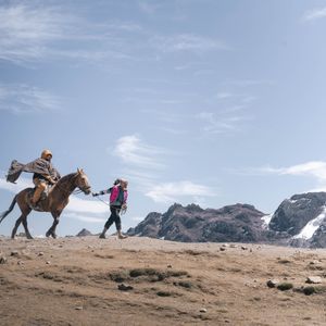 Eine Frau einer WeRoad-Gruppenreise führt ein Pferd mit einem Führer in einem Poncho durch eine felsige Hochgebirgslandschaft mit schneebedeckten Bergen.