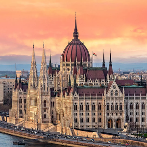 Un edificio del parlamento ornamentado con una gran cúpula se alza a la orilla de un río en el horizonte de una ciudad al atardecer.