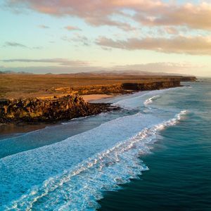 Vue aérienne de vagues turquoise se brisant le long d'un littoral accidenté et aride avec des montagnes lointaines sous un ciel coloré.