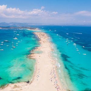 Una vista aerea di una lunga lingua di sabbia che si estende in un mare turchese, con molte barche ancorate e persone sulla spiaggia.