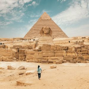 Eine Person läuft in einer sandigen Landschaft unter teilweise bewölktem Himmel auf die Große Sphinx und eine große Pyramide zu.