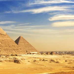 A panoramic view of three large pyramids in a sandy desert with a sprawling city in the distance under a blue, cloudy sky.