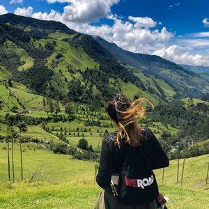 Una mujer con una mochila WeRoad contempla un exuberante valle verde con colinas ondulantes y palmeras altas bajo un cielo parcialmente nublado.