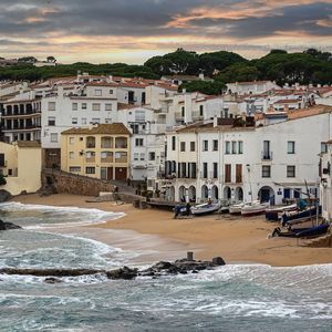 Edifici bianchi di un borgo marinaro si affacciano su una caletta sabbiosa con barche a riva e onde che si infrangono sugli scogli.