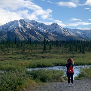 Una donna con uno zaino rosso osserva un fiume davanti a una foresta e una catena montuosa innevata sotto un cielo parzialmente nuvoloso.