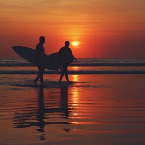Dos surfistas silueteados por el sol poniente llevan sus tablas de surf mientras caminan por la orilla.