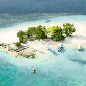 Vue aérienne d'une petite île tropicale avec une plage de sable blanc, des arbres verts et plusieurs bateaux dans l'eau turquoise.