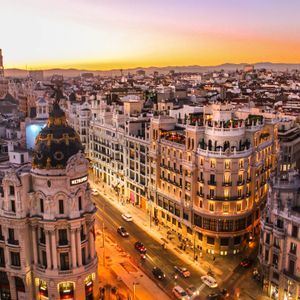 Vista aerea di una fitta città con edifici ornati e strade illuminate al tramonto, con montagne lontane all'orizzonte.