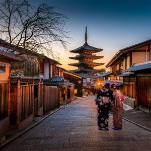 Due donne in kimono tradizionali su una strada acciottolata, di fronte a una pagoda circondata da case di legno al tramonto.